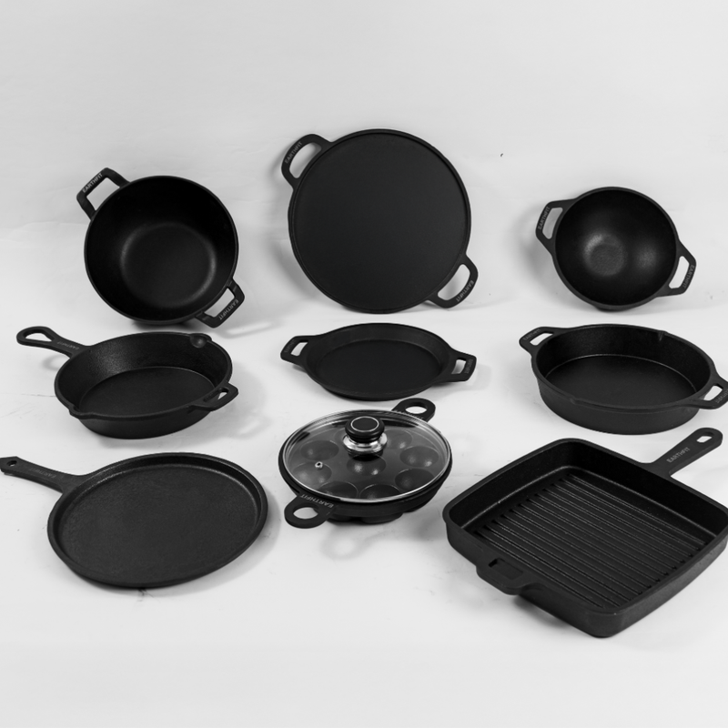 A set of black cast iron cookware pieces arranged on a light gray background, including multiple skillet-style pans, a square grill pan, a lidded pot with a glass cover, and a round lid.